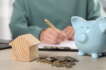 Woman planning budget at wooden table, focus on house model, piggy bank and coins