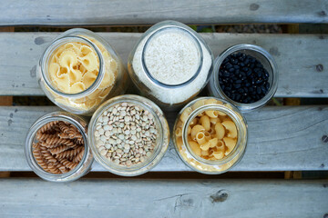 A top-down view of six glass jars filled with various dry foods, including pasta, rice, and beans, neatly arranged on a wooden surface