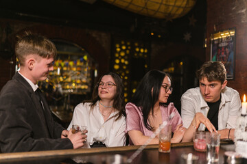 Pretty women with young men wait for orders near bar counter. Friends group has fun with strong...