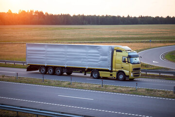 A truck with a tilt semi-trailer carries cargo along the highway in the evening against the...