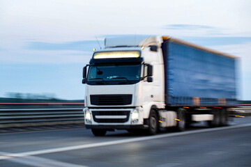 A modern truck with a semi-trailer and headlights on transports cargo in the evening, in the summer, against the backdrop of a field. Trade turnover between countries. Import and export. Blurred