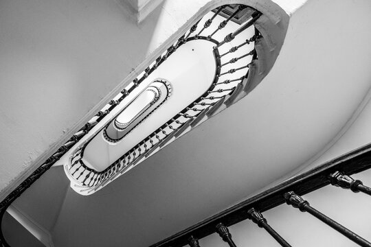 A captivating black and white image capturing the intricate design of a spiral staircase, emphasizing its elongated oval shape and detailed railing. The perspective offers a mesmerizing view that