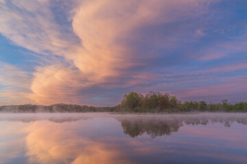 Fototapeta na wymiar Foggy spring landscape at dawn of the shoreline of Whitford Lake with mirrored reflections in calm water, Fort Custer State Park, Michigan, USA