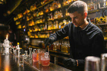 Professional bartender puts ice into glasses with alcoholic beverages using metal tongs. Barkeeper prepares ordered cocktail for serving