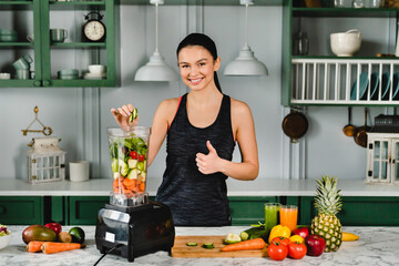Smiling fit woman showing thumb up making green detox juice using blender in the kitchen. Female athlete slimming, drinking super food cocktail and smoothie to burn calories - Powered by Adobe