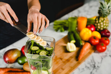 Woman adding banana to the blender with detox green cocktail in the kitchen. Vitamin and fiber...