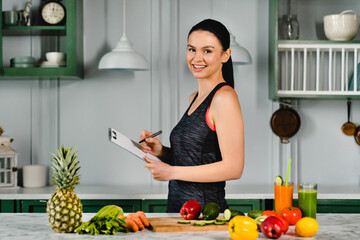 Young slim girl is ready for cooking organic vegetarian food standing with clipboard at the kitchen table, writing counting calories for diet and slimming