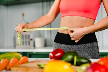 Healthy fit girl measuring her waist with measuring tape in the kitchen. Female athlete eating healthy food, being on a diet for calories burning, losing weight, body shaping