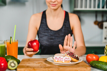 Young fit girl refuses eating doughnuts and opts for healthy food in the kitchen. Difficult choice...