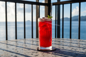 A red cocktail in a tall glass on a table with the sun shining through it. Horizontal picture. 