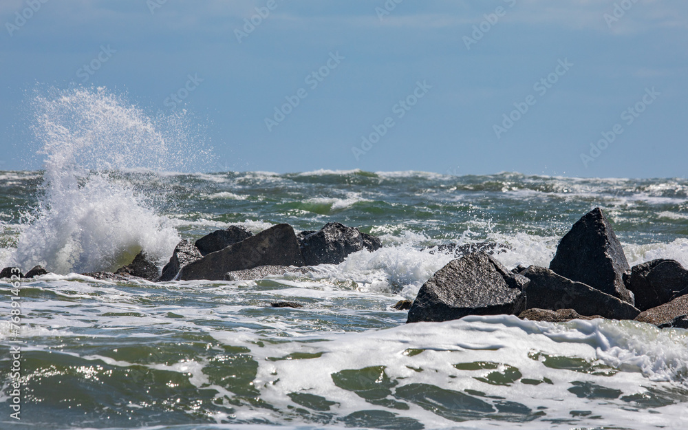 Wall mural Waves crash on dark rocks with a uniform blue sky above.  White Sea foam and ocean spray and small waves in the foreground.  Waves crash during high tide in Florida on the Atlantic Ocean coastline.