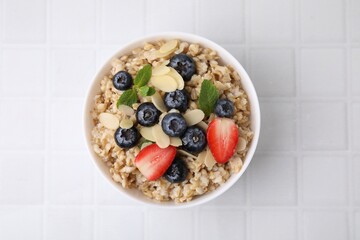 Tasty oatmeal with strawberries, blueberries and almond petals in bowl on white tiled table, top view