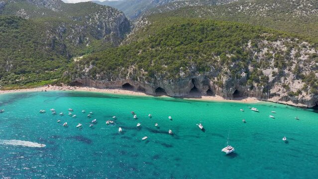 Drone shot - A bay of turquoise blue water full of small boats, visiting a beautiful sandy beach at Cala Luna on the coastline of Sardinia, Italy