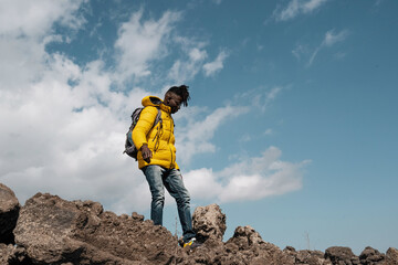 Young African man with vibrant yellow jacket - hiking rocky landscape, adventure fashion - clear...