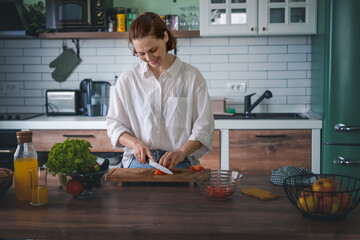 Young Caucasian woman cooking salad from green fresh vegetables while standing in the kitchen at home