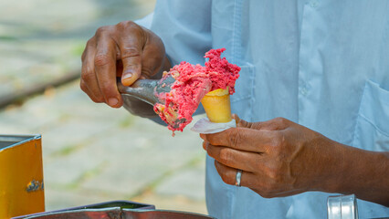 Street vendor in the city preparing and selling a traditional ice cream. Ice cream man serving a scoop of handmade strawberry flavored ice cream in a cone. Typical traditional street ice cream stand.