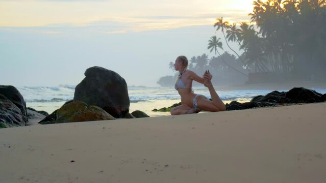 Flexible woman performs leg pigeon pose on shore with palm trees. Young lady enjoys yoga in relaxing atmosphere of ocean