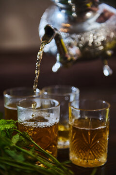 Pouring delicious Moroccan mint tea from a silver teapot into drinking glasses. Bunch of fresh mint in the foreground