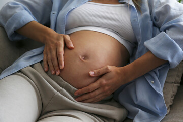 Young woman on a third trimester of pregnancy. Pregnant female sitting on the couch with arms on her belly. Expecting a child concept. Background, copy space.