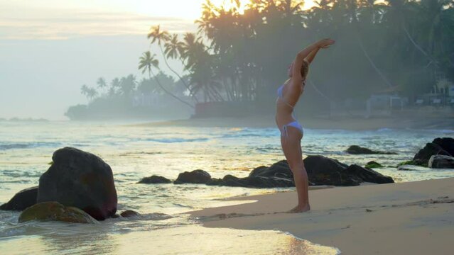 Young woman in bikini performs tadasana asana standing on beach. Female athlete engages in yoga enjoying tropical view at sunset