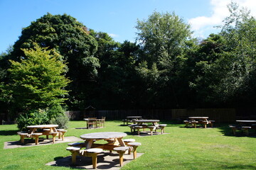 Typical British countryside pub beer garden. Image taken before opening with no customers. Wooden tables scattered across the green lawn. A forest in the background with a cloudy sky.  - obrazy, fototapety, plakaty