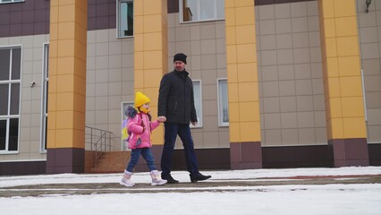 little child with backpack goes school, holding his father hand, caring young father, happy family, cheerful child girl walks with backpack into school building, snowy weather, people outdoors winter