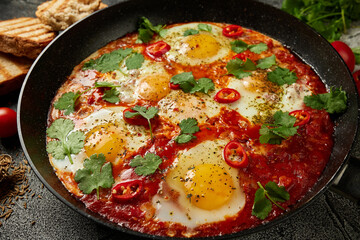 Tasty Shakshuka eggs in a pan with toast on a dark concrete background. Eggs poached  in a spicy tomato pepper sauce. Traditional Jewish scrambled eggs.