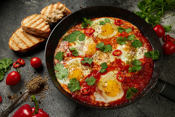 Tasty Shakshuka eggs in a pan with toast on a dark concrete background. Eggs poached  in a spicy tomato pepper sauce. Traditional Jewish scrambled eggs.