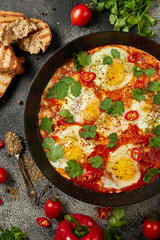 Tasty Shakshuka eggs in a pan with toast on a dark concrete background. Eggs poached  in a spicy tomato pepper sauce. Traditional Jewish scrambled eggs. Flat lay, Top view.