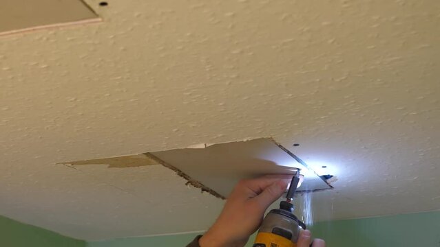 Man fixing hole in the ceiling with patch or piece of plasterboard or drywall using electric cordless drill screwdriver.
