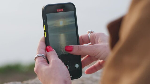Close-up video of gentle woman's hands, adorned with red nails, tapping on smartphone screen, capturing sunset on a beach.