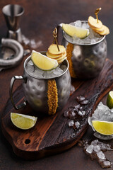 Two silver mugs of moscow mule cocktail decorated with cane sugar, lime and slices of ginger on wooden board. Side view, close up
