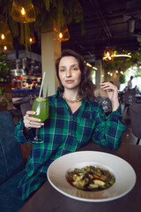Portrait of a woman in a shirt sitting in a restaurant with a salad and a green smoothie held in her hands.