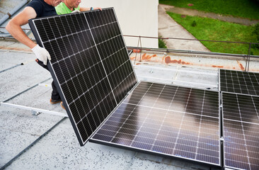 Workers building solar panel system on metal rooftop of house. Two men installers installing photovoltaic solar module outdoors. Alternative, green and renewable energy generation concept.
