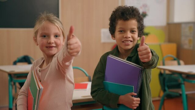 Two happy pupils children diverse multiethnic kids African American boy and Caucasian girl classmates friends showing thumbs up likes recommend gesture at school class holding books study good learn