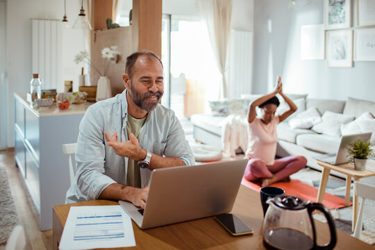 Man working on laptop with woman in background doing yoga at home - Powered by Adobe