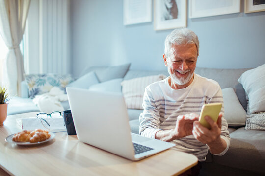 Senior man using smartphone at laptop on home sofa