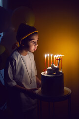 portrait boy in a yellow hat and white T-shirt blows out candles from a cake with a goose in the dark.
