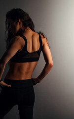 Female sport muscular woman posing in black sport bra showing the shoulders, blades and arms, standing on studio wall background with empty copy space. Back view. - 769057274