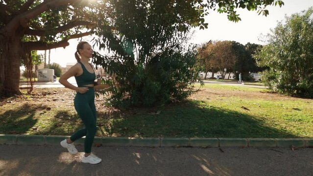 A beautiful fair-haired European woman goes for a run in a green city park on an early summer morning. Sunlight filters through the foliage, and the camera moves alongside her. Slow-motion