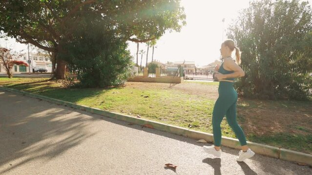 On a bright  summer morning, a stunning fair-haired European woman goes for a jog in a lush green city park, shaded by trees. She moves gracefully from right to left as camera captures in slow motion