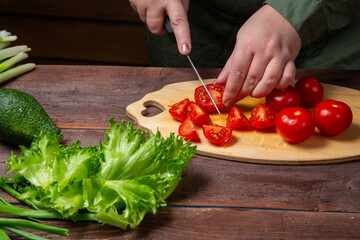 A woman's hand cuts a cherry tomato with a knife on a board on a table with vegetables