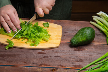 A woman's hand cuts greens in half with a knife on a wooden board on a table with vegetables
