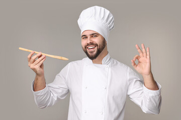 Happy young chef in uniform holding wooden spoon and showing ok gesture on grey background