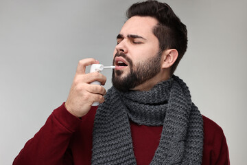 Young man with scarf using throat spray on grey background