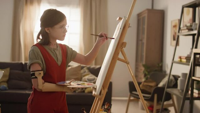 Side full medium footage of Caucasian young teenage girl in jumpsuit painting on canvas while holding color palette with bionic arm at home alone on sunny day