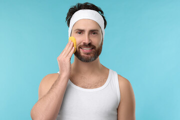 Man with headband washing his face using sponge on light blue background