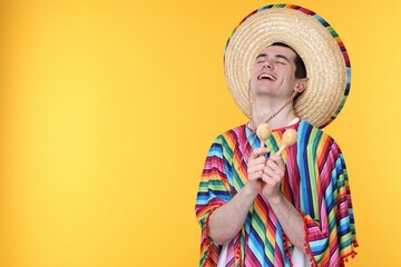 Young man in Mexican sombrero hat and poncho with maracas on yellow background. Space for text
