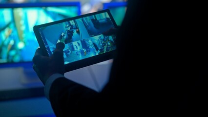 Man security guard officer working on tablet in monitoring center, zooming watching video. Computers and big digital screen show cctv cameras footage.
