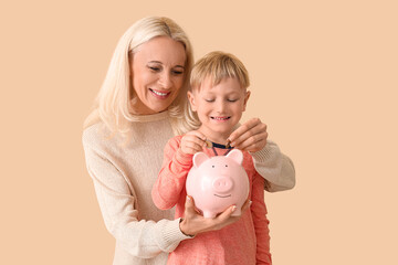 Little boy with his grandmother putting coins in piggy bank on beige background